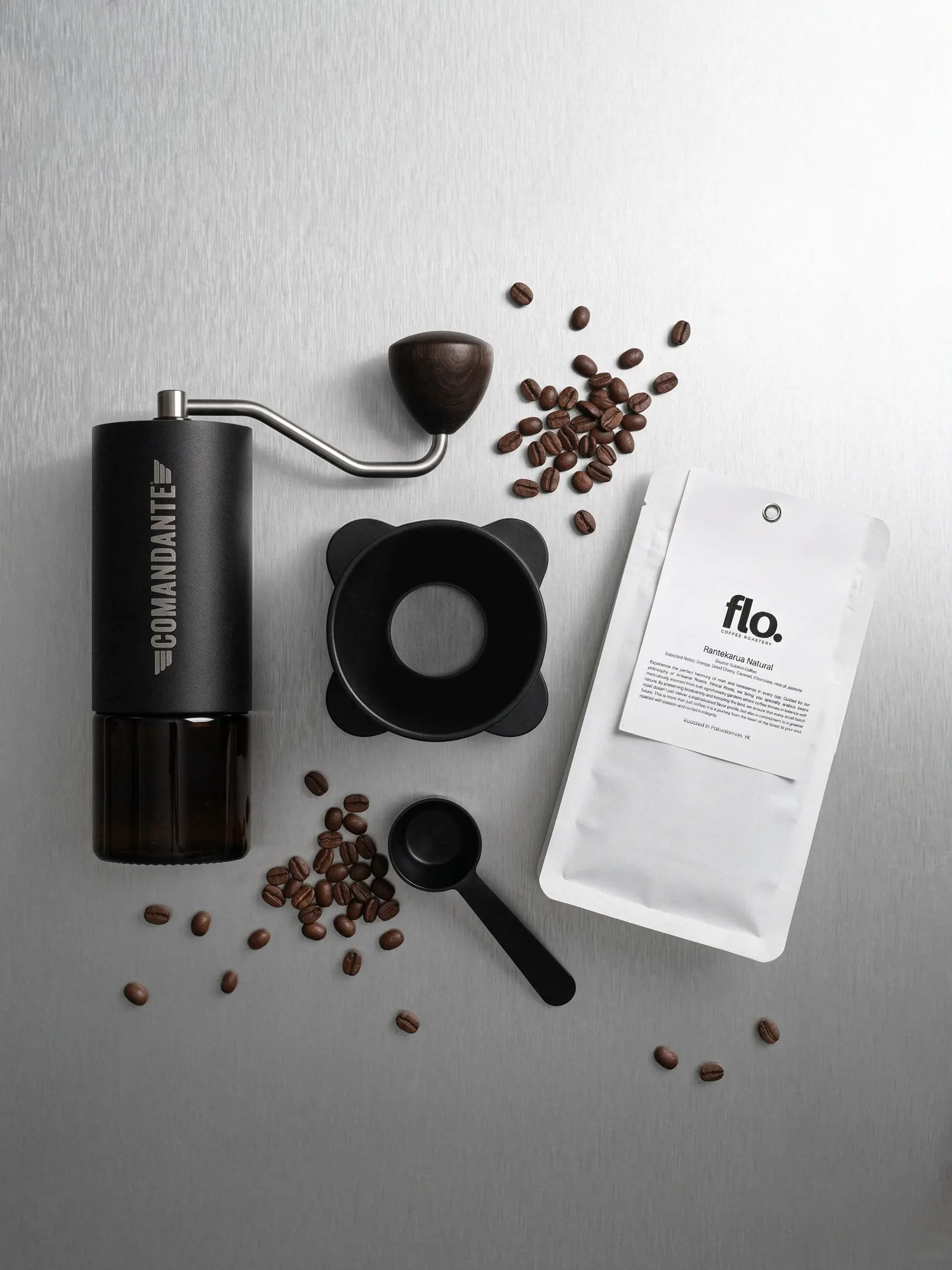A hand grinder, a dosing funnel and a bag of Rantekarua Natural on brushed steel, with roasted beans scattered around.